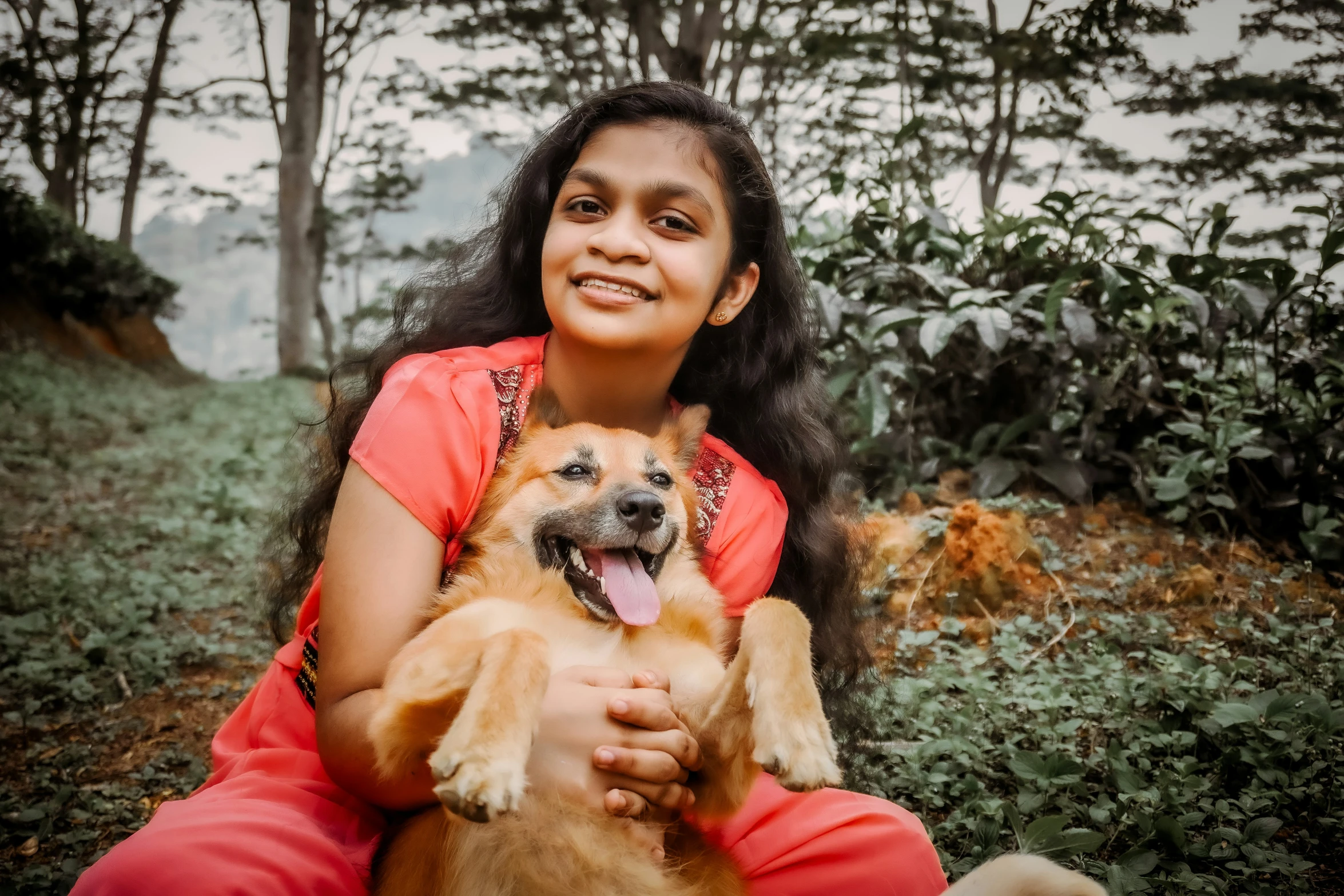 a woman sitting in the woods holding a brown dog