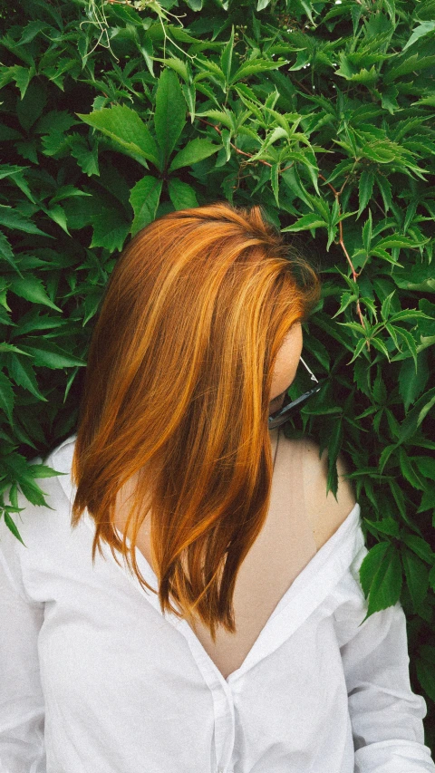 a woman in a white top stands with her back to the camera with leaves and plants behind her