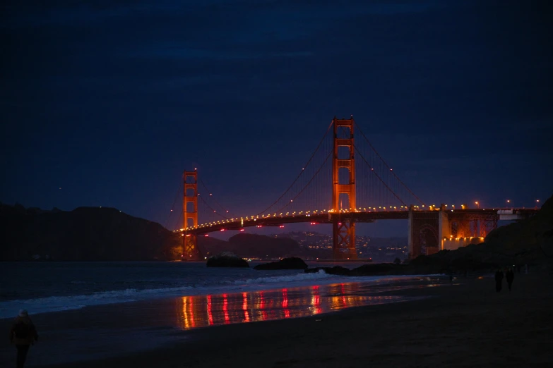 the lights of a bridge shine on a beach as people walk past