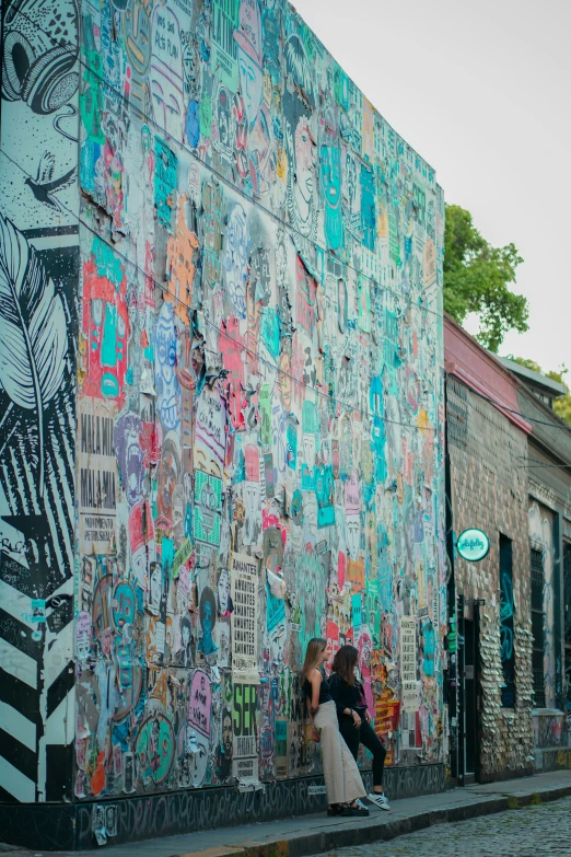 two people standing on the sidewalk in front of a wall that has various colored stickers all over it