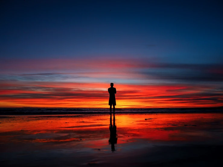 a silhouetted person standing on the beach as the sun sets