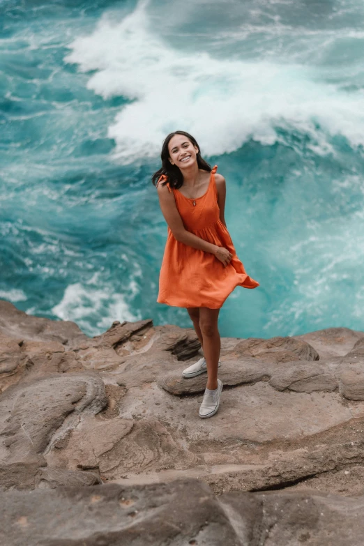 the woman in an orange dress is standing on a rocky shoreline near a wave