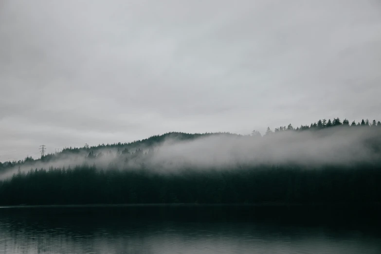 a foggy lake on a dark day, with trees on a mountain in the distance