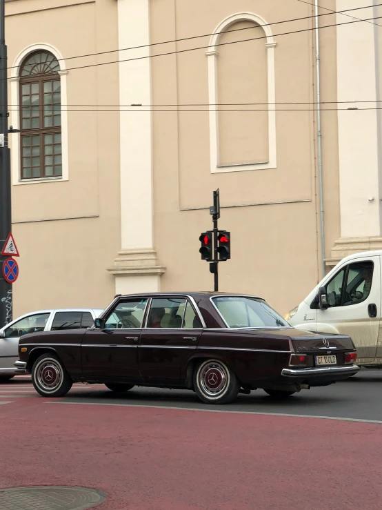 a classic car parked on a side walk as traffic crosses in front of a building