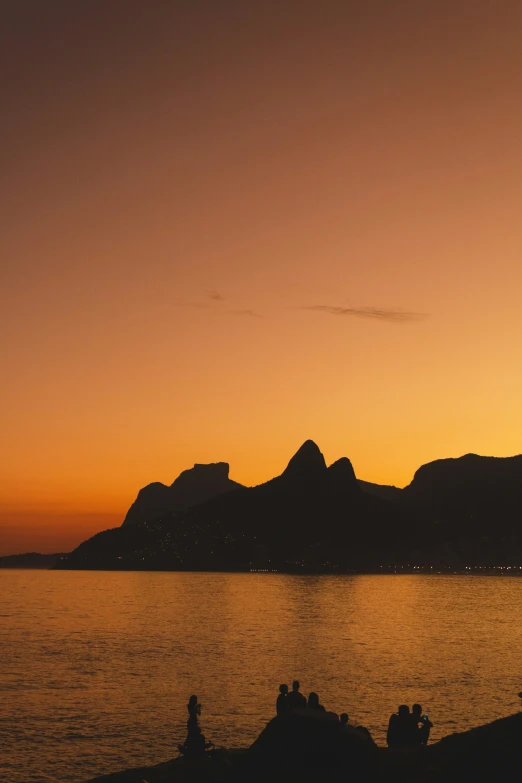 the sun setting over the water and people sitting on rocks