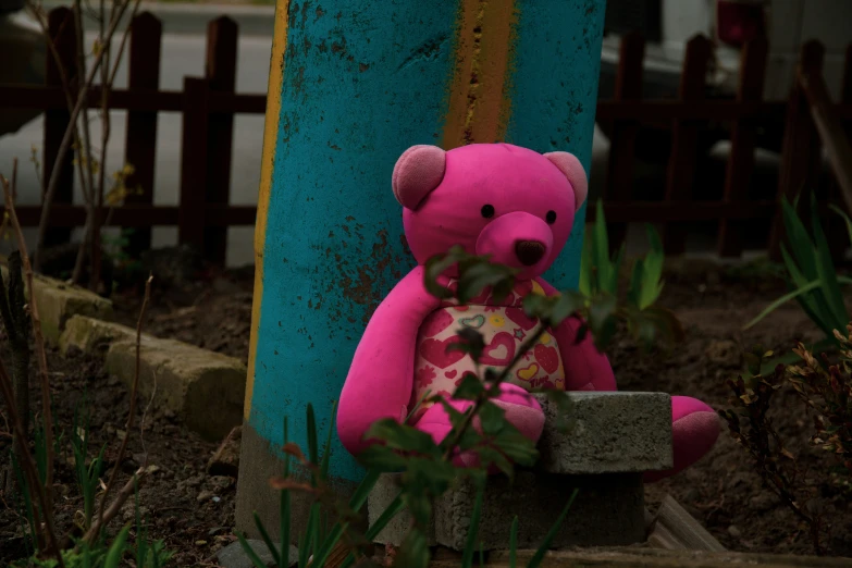 a pink teddy bear sitting in the bushes next to a tall wooden pole