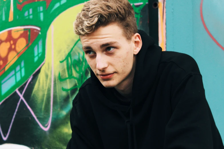 a man in black hoodie sitting next to a colorful wall