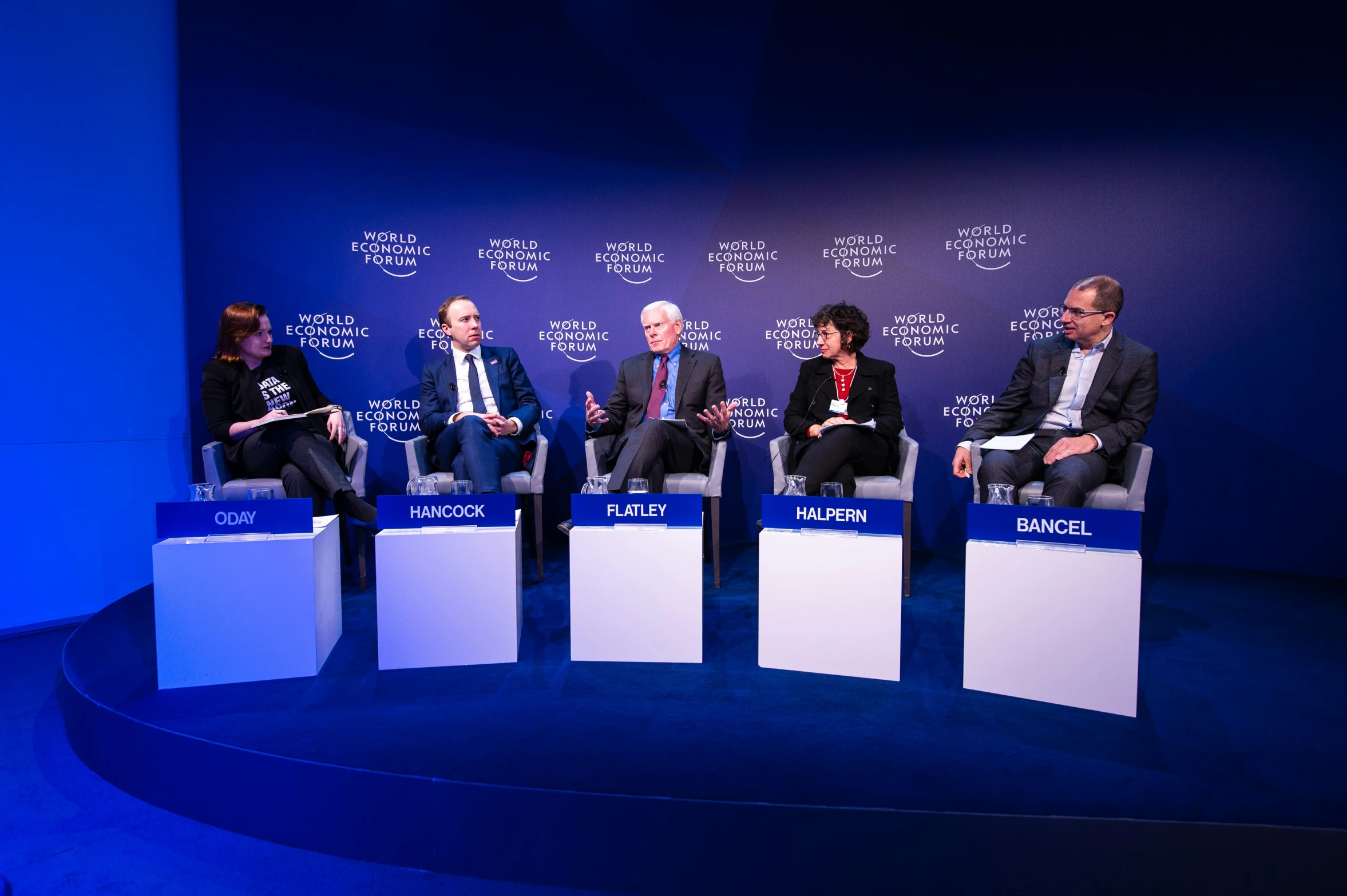 four people in suits sitting at tables on blue lighting stage