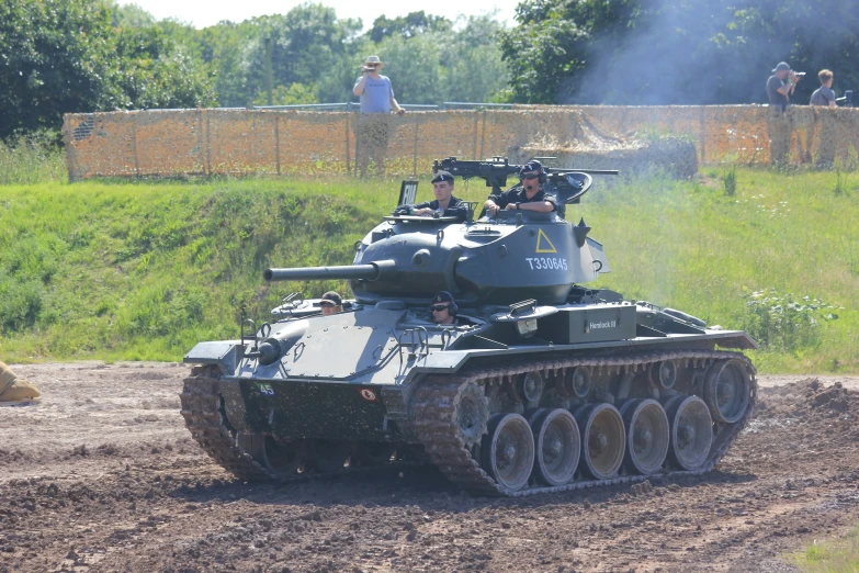 an army tank driving on dirt with some people watching