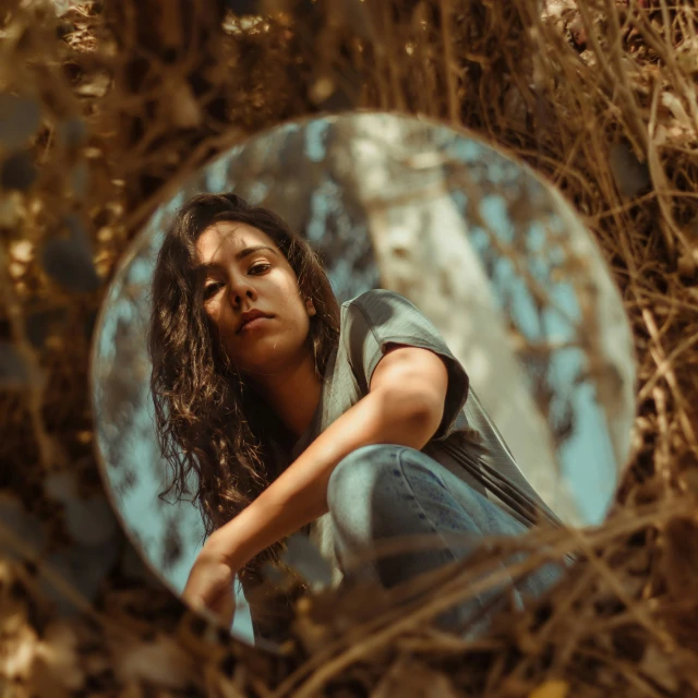 a woman sitting in dried grass and looking in a mirror