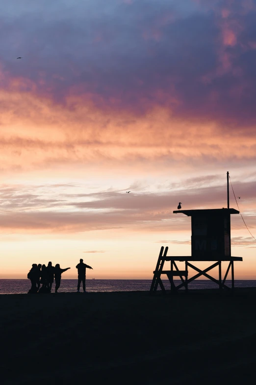 people are standing on the beach near a lifeguard tower at sunset