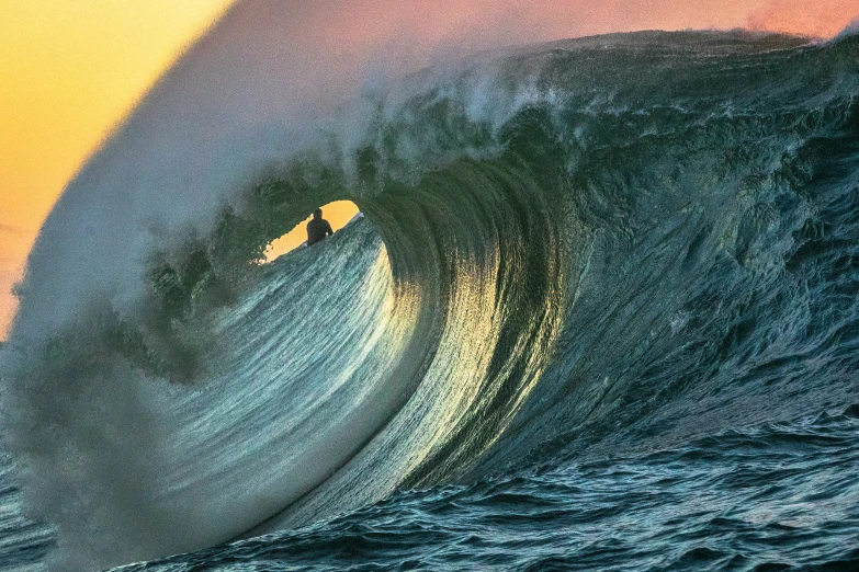a large wave breaks into the sunset in front of a surfer
