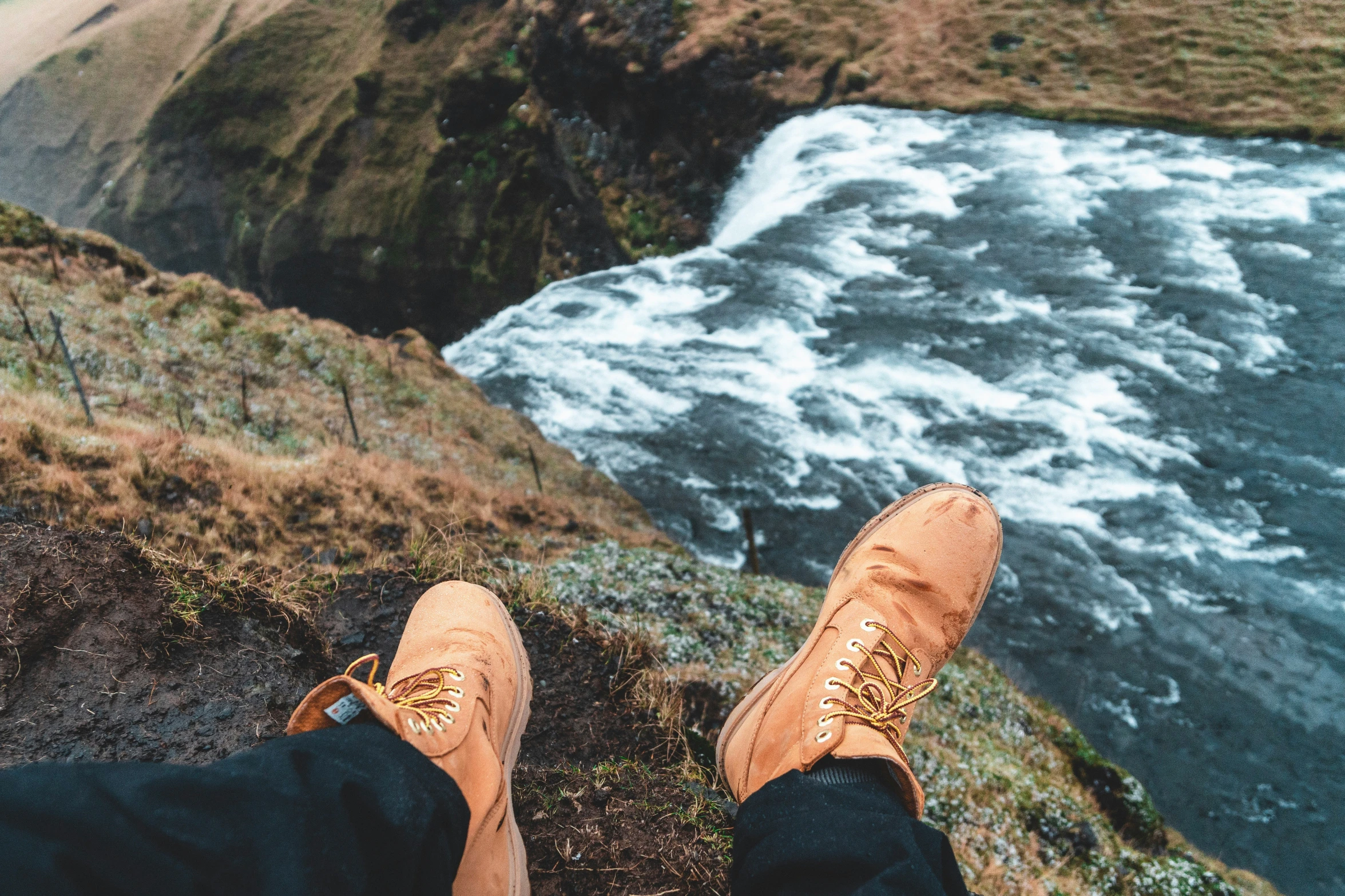 someone wearing brown shoes standing at the edge of a cliff with a river and a canyon in the background