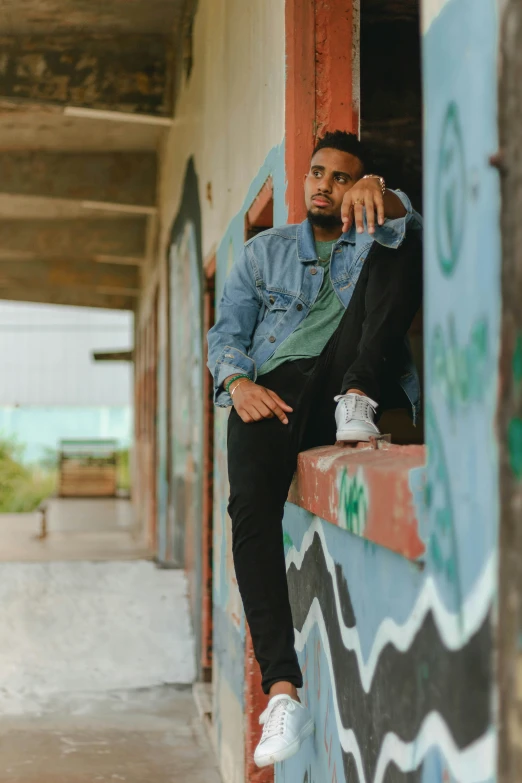 a black guy wearing a blue jean jacket sits on the outside of a building