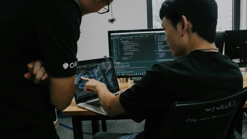 two men in a computer lab looking at a laptop and another works on his computer