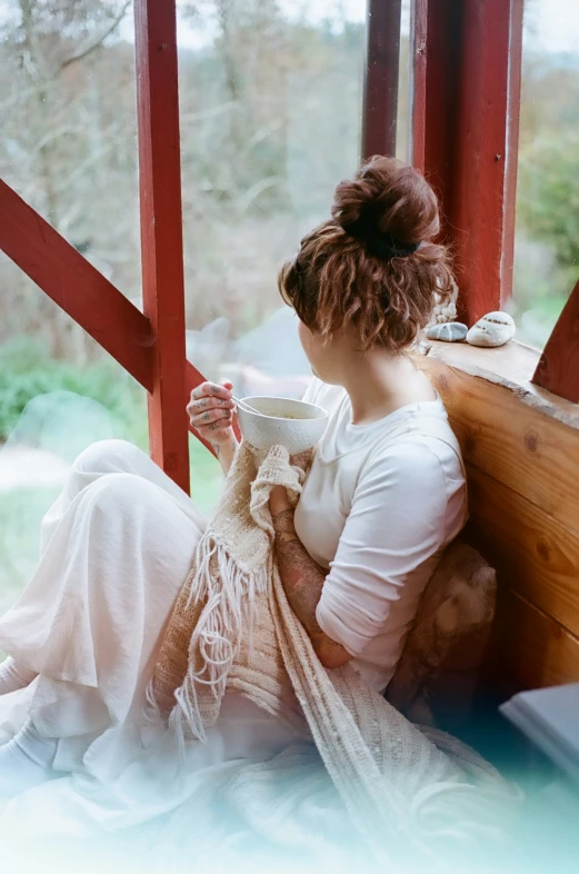 a woman sitting down in front of a window drinking from a cup