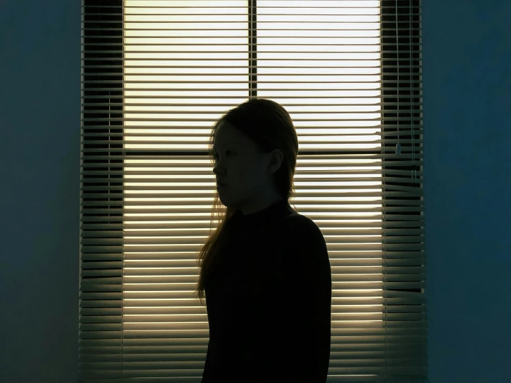 a girl stands in front of a window with blinds