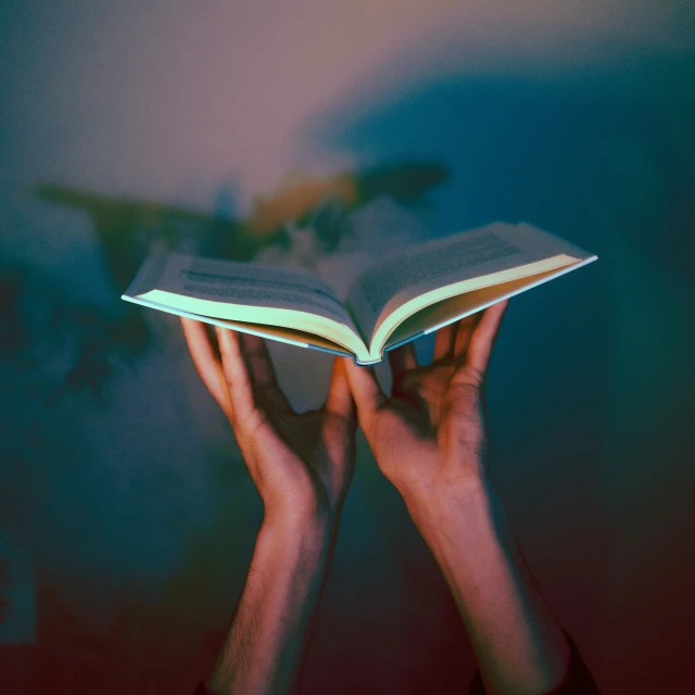 two hands holding an open book with dark paint and lighting from the bottom