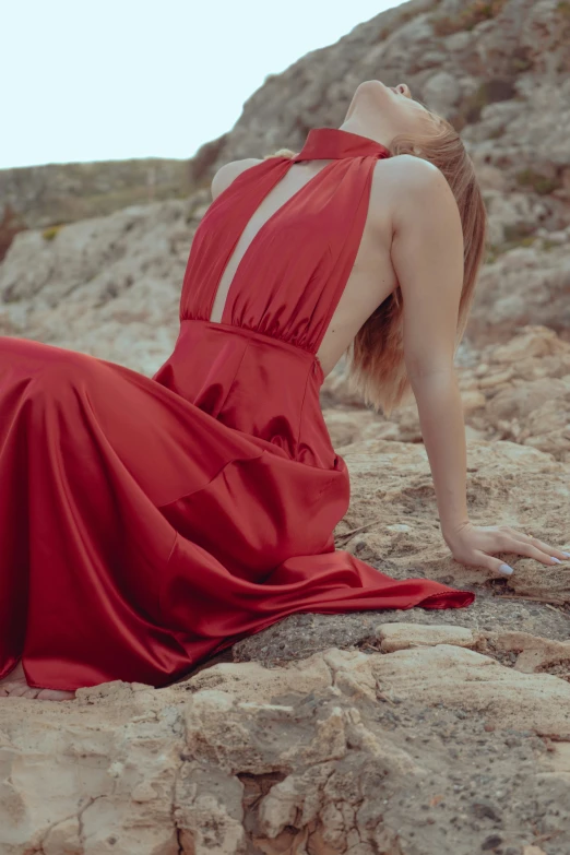 a woman with long hair, dressed in a red gown sits on rocks and leans in her left arm