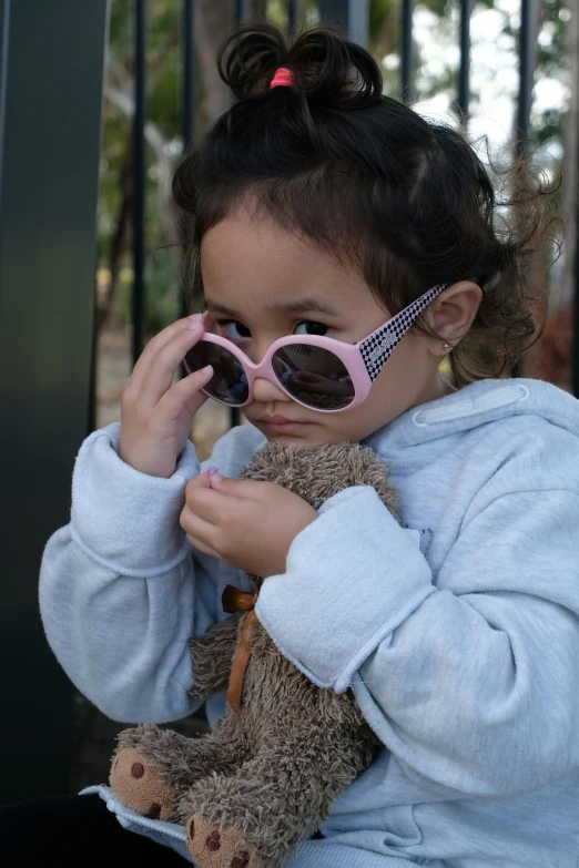 a little girl with pink sunglasses sitting down holding a teddy bear