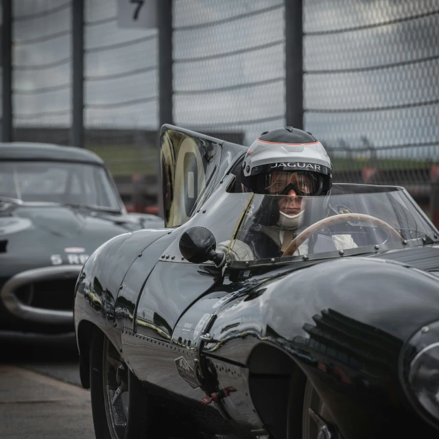 a woman in a vintage sports car has her hand up to the driver's head