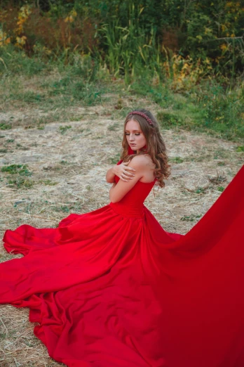 a girl with long hair and wearing a red dress sitting in some brown grass