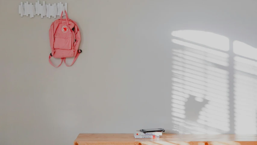 the pink backpack hangs on the wall next to a window with blinds