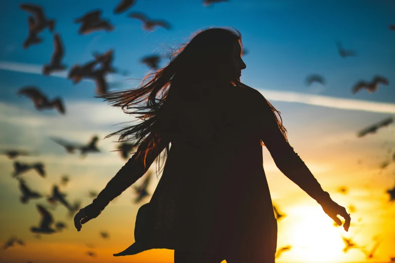 a woman standing in front of the sunset with birds flying around her