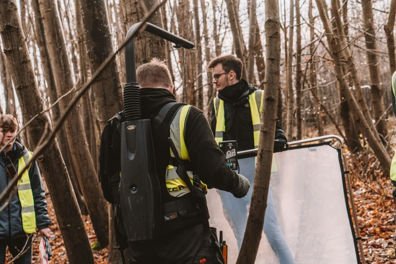 two men in black jackets and reflective vests stand in a wooded area, one of them holds a large mirror