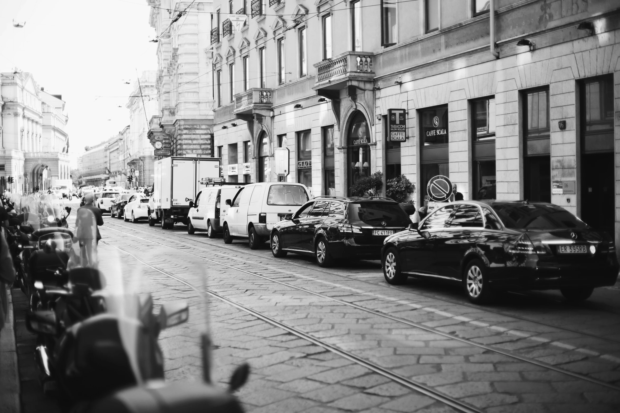 cars line a street as pedestrians walk by in front of tall buildings