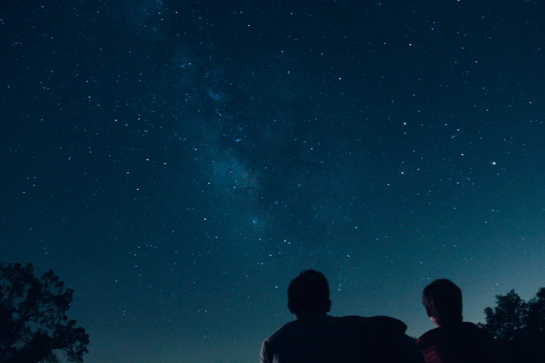 two people sit near a tree on the edge of a hill and look at the sky, filled with stars