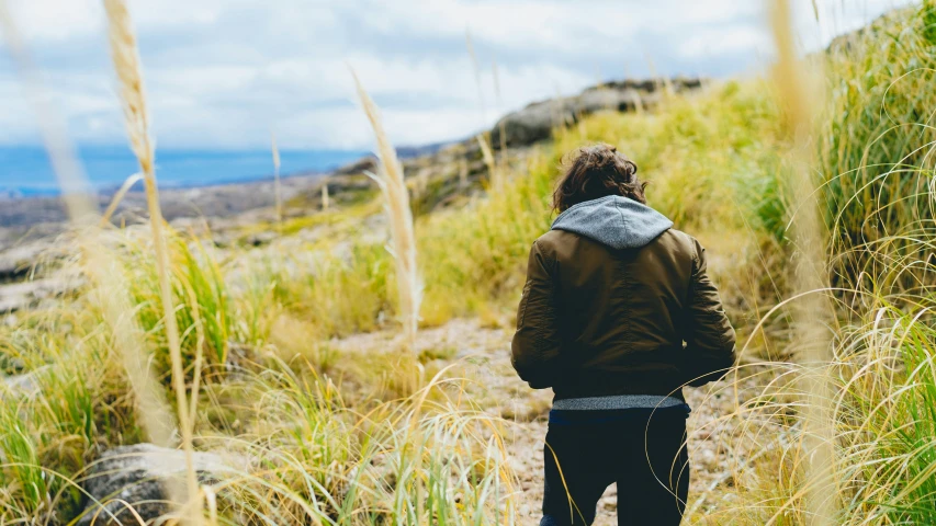 a man in a parka walking up a hill with tall grass and grasses