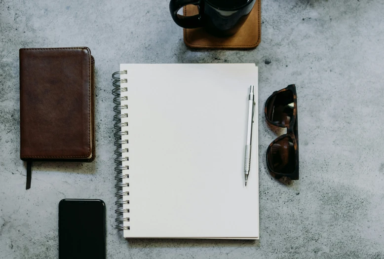 a notebook and sunglasses, coffee mug, sunglasses and phone sit on the cement ground