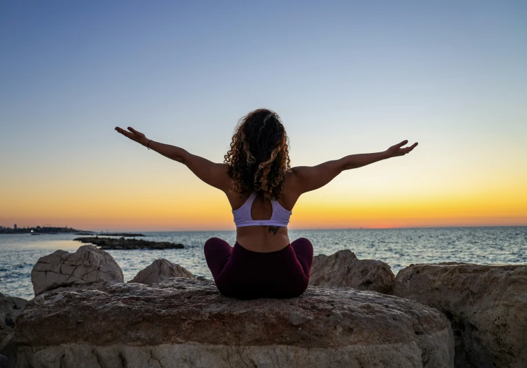 a woman sitting on the rocks, with her arms outstretched as she looks out at a beautiful ocean