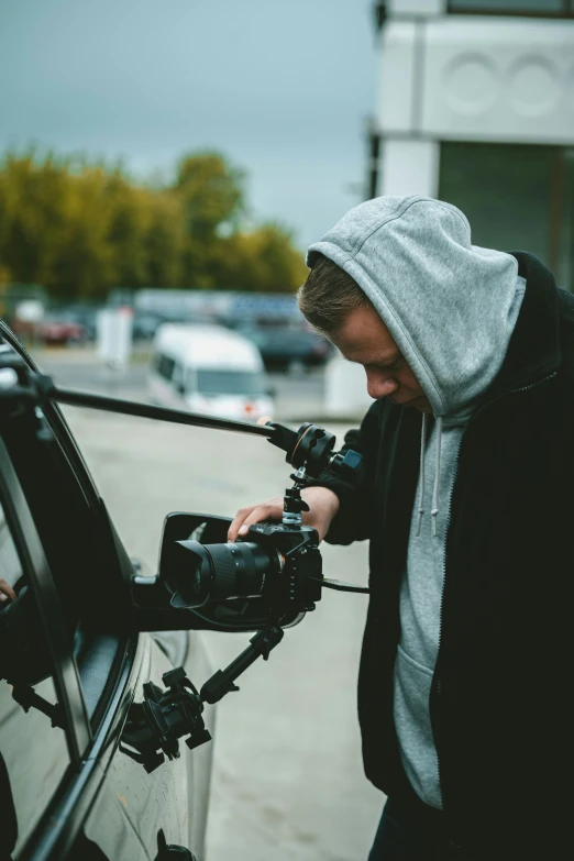 a man with a hoodie and holding a camera by a car