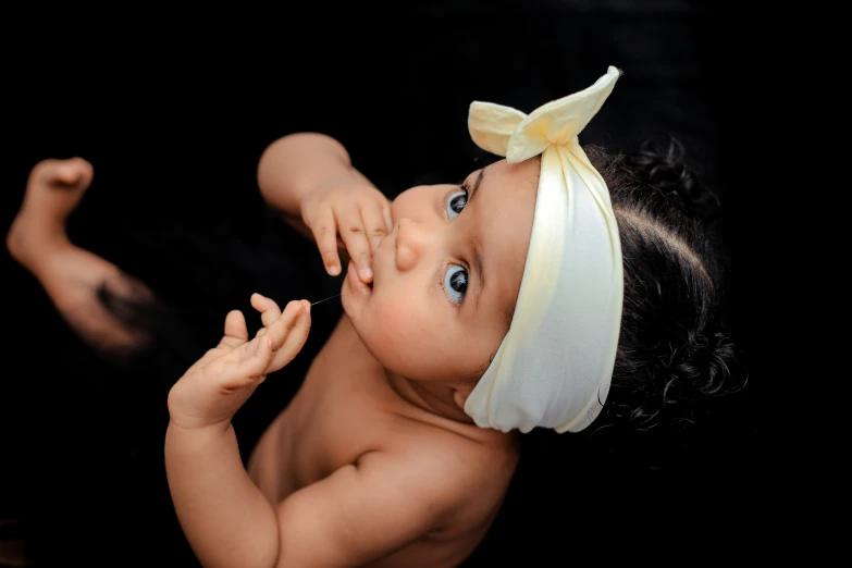 an infant girl with a white headband on holding her hand up to her mouth
