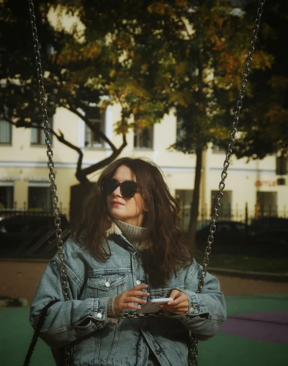 a young woman wearing sunglasses and a jean jacket on a swing