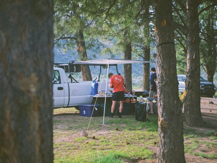 two men standing next to a tent and a van in a forest