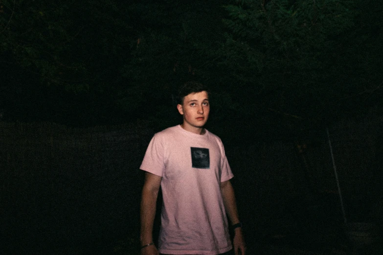 a young man stands in front of dark green trees