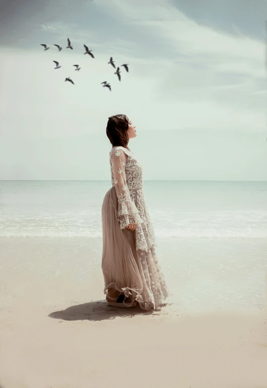 woman in flowing dress standing near ocean and flock of birds