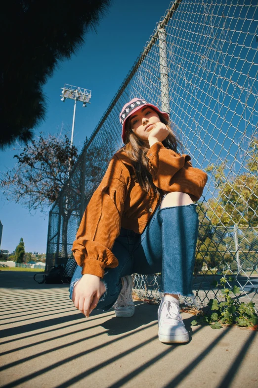  in orange sweater and blue jeans with tennis shoes leaning against fence