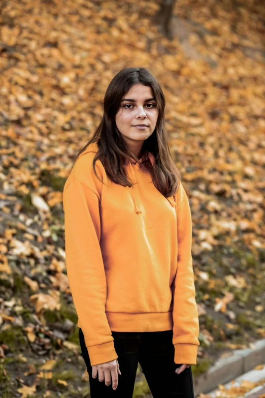 a  in an orange sweatshirt is standing outside with leaves all around her