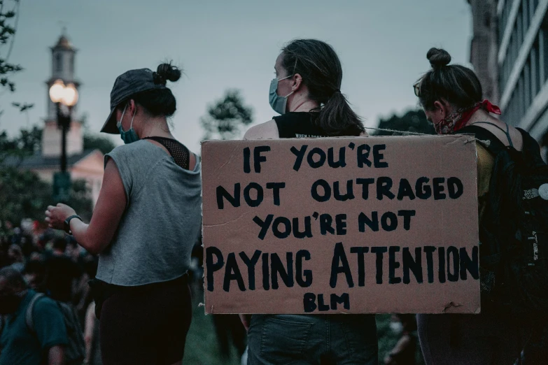 four women holding a sign saying if you're not outaged you're not paying attention