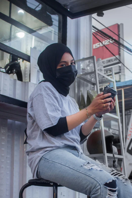 a woman in jeans and face mask on a stool using her cell phone