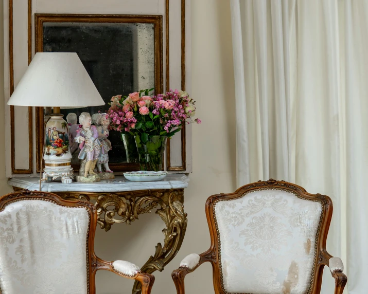 two antique chairs in front of a table with flowers and a vase of flowers