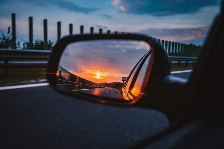 a rear view mirror reflecting the sun going down on the highway