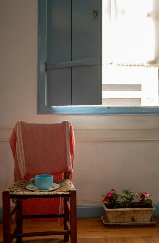 a chair sitting by the window, with an empty tea cup and saucer on a saucer on a stool in front of it