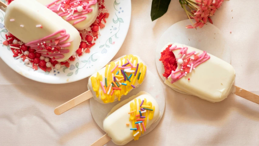 two plates with cake pops on them next to flowers and one plate has red, white, yellow, and blue sprinkles