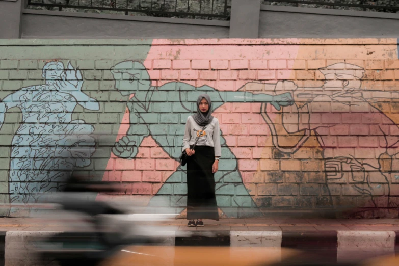 a woman in grey shirt and black pants standing in front of graffiti on a brick wall