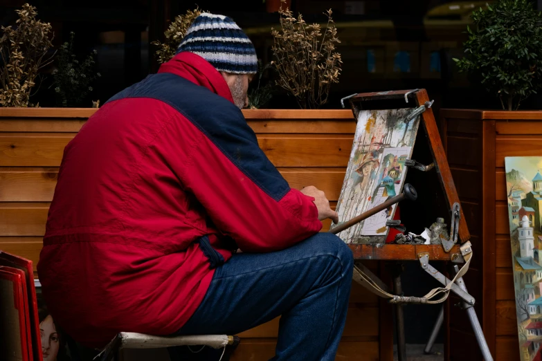 a man in winter clothes sits near a painting on a easel